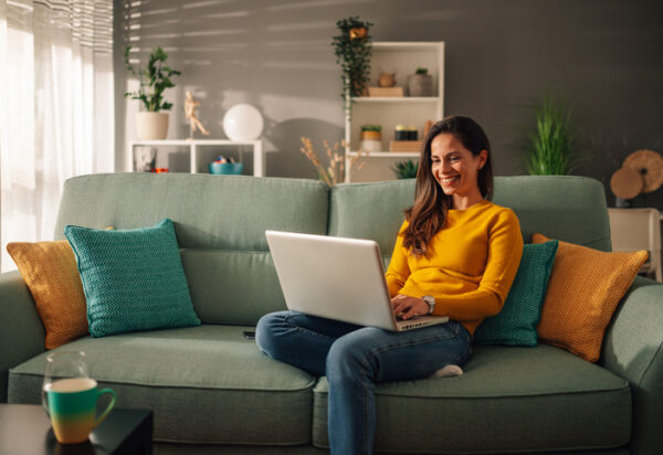 Smiling caucasian woman using laptop while sitting on a mint couch at home. Happy female shopping or chatting online in social network, typing blog, freelancer working on a project.