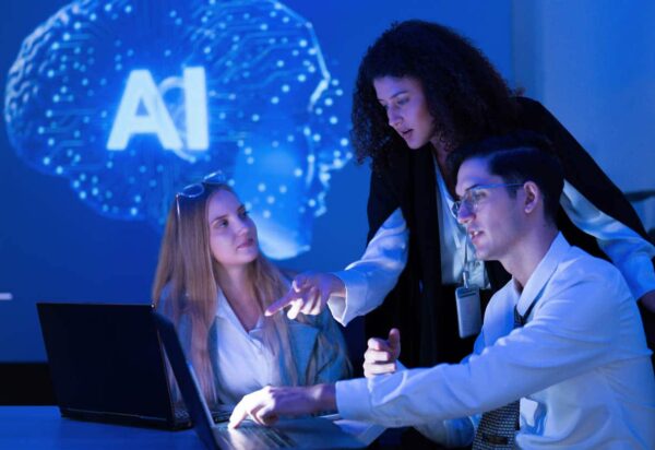 A group of three professionals engages in a discussion about an artificial intelligence project using a laptop. They work in a contemporary setting with blue ambient lighting.